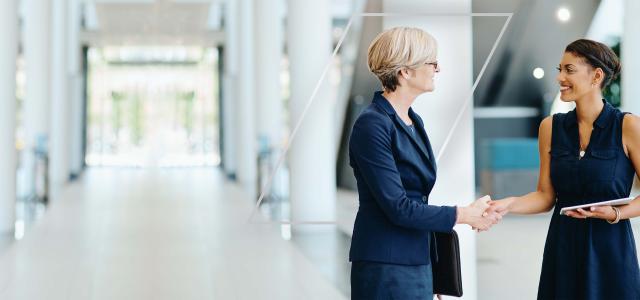 Two women shaking hands and smiling cheerfully while standing in modern office interior